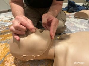 an image of a man's hands pinching a manikin's nose with 2 fingers on its chin, tilting the manikins head back. The manikin is on the carpeted floor of an ofice for CPR certification in Atlanta