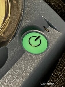 The green, power button of an AED is shown which has the universal power symbol of a circle with a vertical line at it's 12 o'clock position.